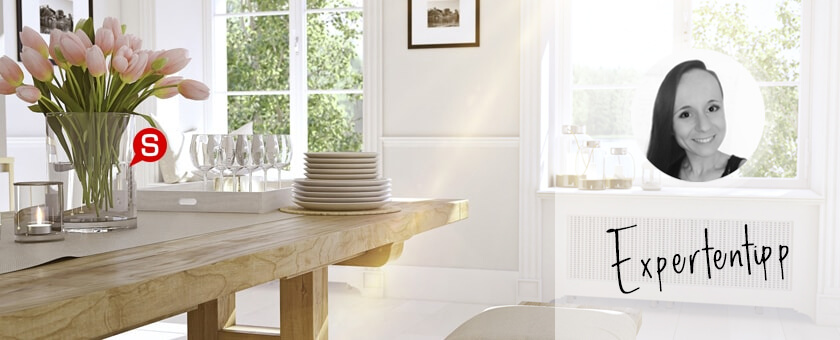 Ein helles Esszimmer im skandinavischen Einrichtungsstil mit einem Esstisch aus Holz. Auf dem Tisch befindet sich eine Glasvase mit Blumen. Das Gesamtbild wird von Bildern in schwarzen Rahmen an weißen Wänden abgerundet.