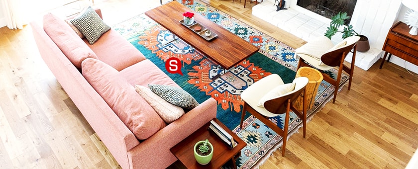 Ein Wohnzimmer im Boho-Stil mit einem bunten Teppich. Auf dem Teppich steht ein rosafarbenes Sofa mit Dekokissen und ein holziger Couchtisch mit Dekorationen.