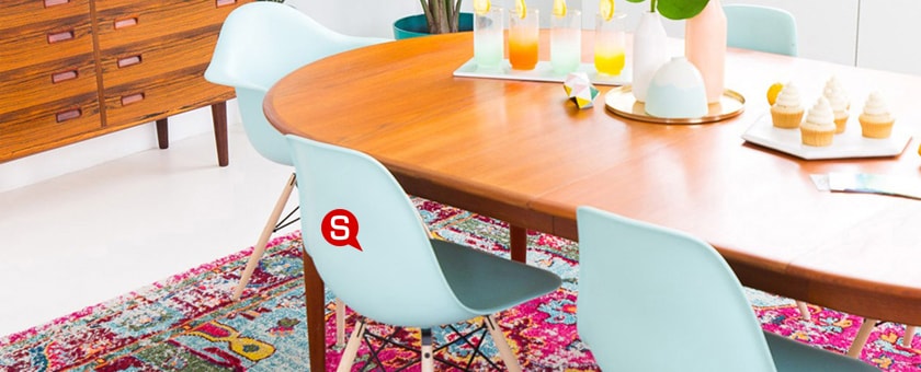 Ein Esszimmer mit einem großen Tisch in Holzoptik, an dem hellblaue Kunststoffstühle stehen. Die Stühle kontrastieren mit zarten rosafarben und weißen Blumenvasen auf dem Tisch. Unter dem Tisch liegt ein bunter, alt gemachter Teppich. Weiße Wände mit weißem Boden runden das Gesamtbild ab.