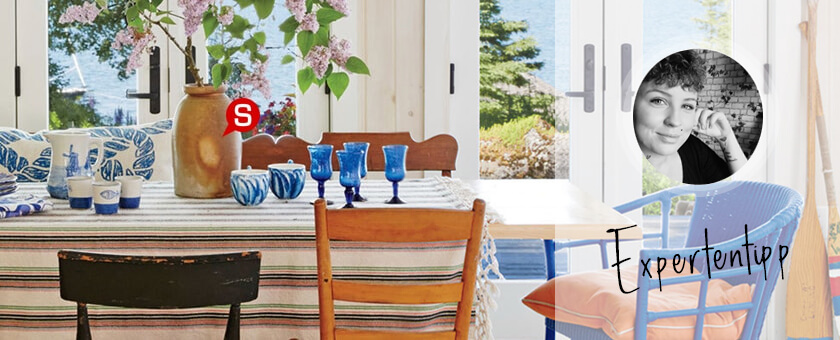 Ein helles Esszimmer im Boho Style mit einem großen Esstisch aus Holz, an dem Tisch stehen alt gemachte Holzstühle verschiedener Form und einem blauen Rattansessel. Auf der Tischplatte stehen eine Vase mit Blumen und jede Menge Dekorationen in Weiß und Marineblau.