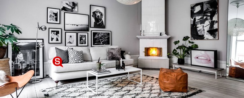 Ein Wohnzimmer im skandinavischen Stil mit grauen Wänden und einem hellem Sofa. Auf dem Sofa liegen Dekokissen und darüber hängen schwarzweißen Bilder. Vor dem Sofa steht ein weißer Couchtisch mit silberner Lampe.